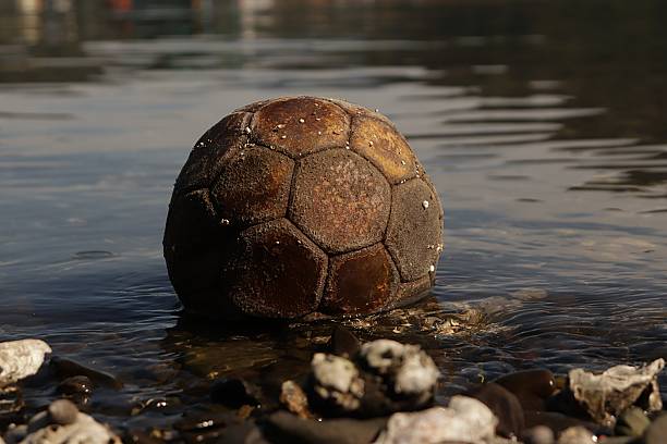 A heavily weathered and dirty brown soccer ball, showing signs of significant wear and tear, floats gently on the calm, clear surface of shallow water near a rocky shore, symbolizing themes of abandonment, forgotten childhood, and the passage of time.