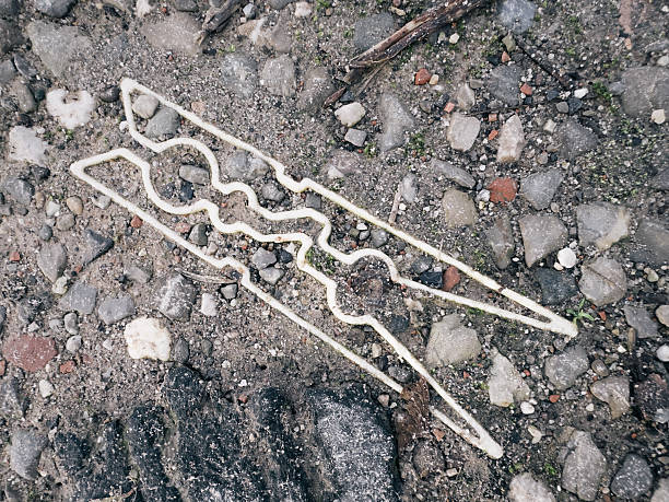 An overhead view of a white, clothespin-shaped form partially embedded in rough gravel and soil. The object appears flat and integrated into the ground surface, blending an everyday household shape with an outdoor, weathered environment. The image presents a visual ambiguity between found object, outline, and installation-like intervention, emphasizing contrast between artificial form and natural textures.