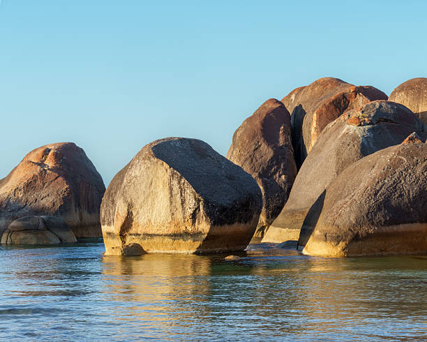 Rounded granite boulders rise from shallow coastal water under clear, warm light. The scene highlights the distinctive formations of a well-known coastal area in Western Australia.