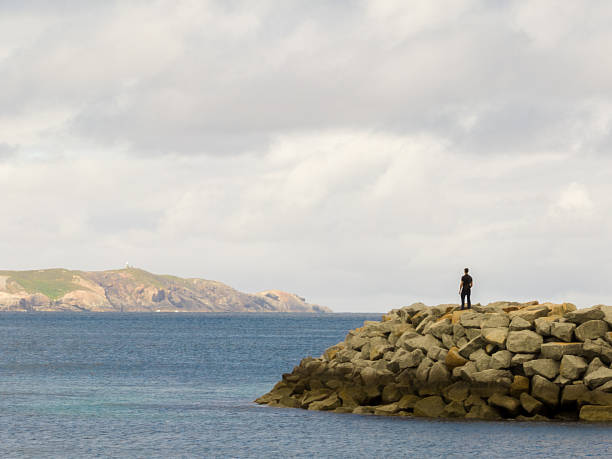 A distant figure stands on a rocky breakwater overlooking calm blue water with hills visible in the background. The scene shows a coastal structure and surrounding seascape under soft daylight.
