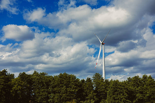 A tall wind turbine is visible among lush green trees, set against a backdrop of a clear blue sky filled with large white clouds, indicating a bright day.