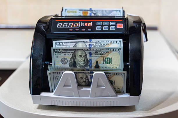 istock Automatic machine counting stack of US dollars. Front view of a digital money counter processing 100 US bills, showing display and control panel on a table indoors. 2254341751