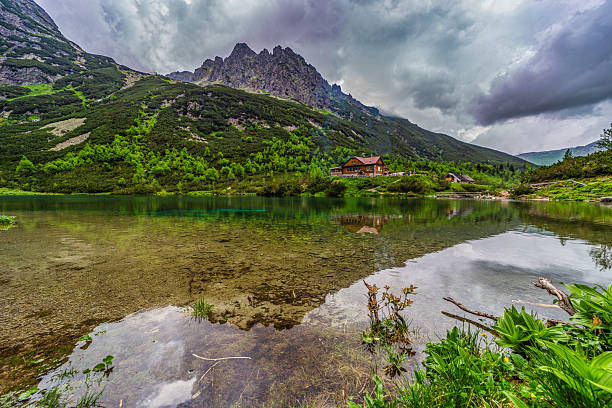 A dramatic landscape shot of a hiking path at the edge of Green Lake (Zelene pleso). Massive granite peaks with snow patches are shrouded in heavy grey storm clouds. Tatra National Park, Slovakia.
