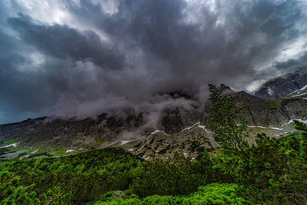A dark and moody mountain landscape in the High Tatras, Slovakia. Heavy storm clouds and thick fog descend upon the jagged granite peaks and green slopes, creating a powerful atmosphere of wild nature.