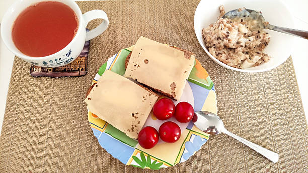 Healthy breakfast is served on a table with place mat, including fruit tea, cheese sandwiches, cherry tomatoes and oatmeal with milk and flax seeds