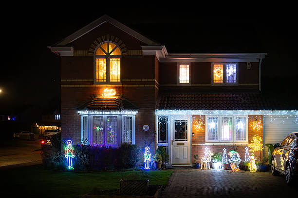 A two-story suburban residence featuring brick and white siding is elaborately decorated for the Christmas holiday season. Multicolored string lights outline the roofline, windows, and doorway, creating a festive glow against the dark night sky. Two illuminated nutcracker figures, one blue and one green, stand sentry on the front lawn amid evergreen shrubs. A large wreath with lights adorns the front door, flanked by potted plants and additional light displays. Warm light emanates from several windows, suggesting interior holiday preparations. A driveway leads to a two-car garage, with two vehicles parked nearby. The scene is set outdoors in a residential neighborhood during winter, emphasizing traditional holiday decorations. No people are visible in the image.