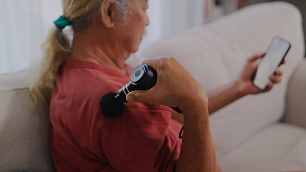 istock Elderly Asian Man Using Massage Gun While Checking Smartphone at Home. 2254354078