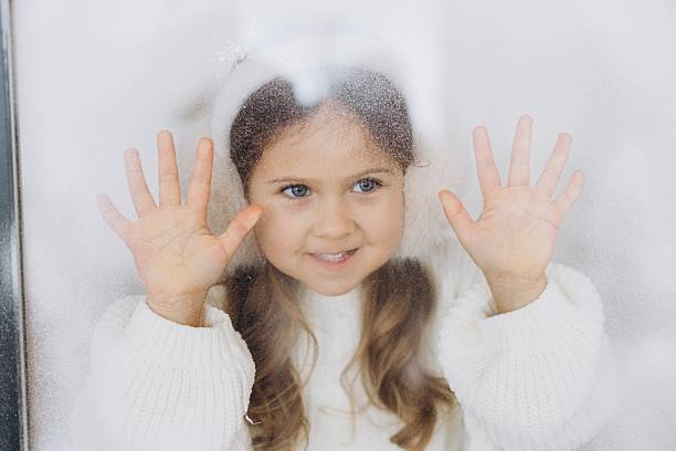 Little girl smiling, pressing hands on a frosted window, looking out at cold winter weather, feeling curious
