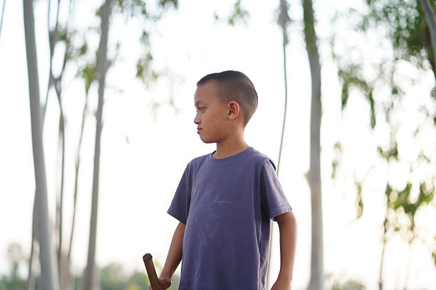 Young boy stands outdoors, surrounded by tall trees, holding stick. His expression is thoughtful, reflecting moment of contemplation in nature