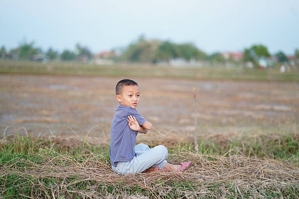 Young boy sits on dry grass in rural landscape, looking back with thoughtful expression. background features vast field, suggesting serene and peaceful environment