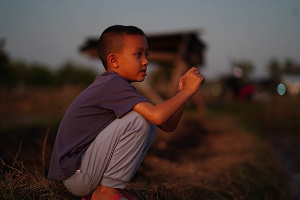 Young boy squats thoughtfully in serene outdoor setting during sunset, surrounded by nature. His expression reflects curiosity and contemplation, capturing moment of peaceful solitude