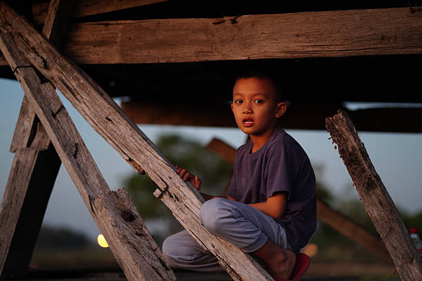 Young boy sits under wooden structure, surrounded by rustic beams. warm light of sunset creates serene atmosphere, highlighting his curious expression