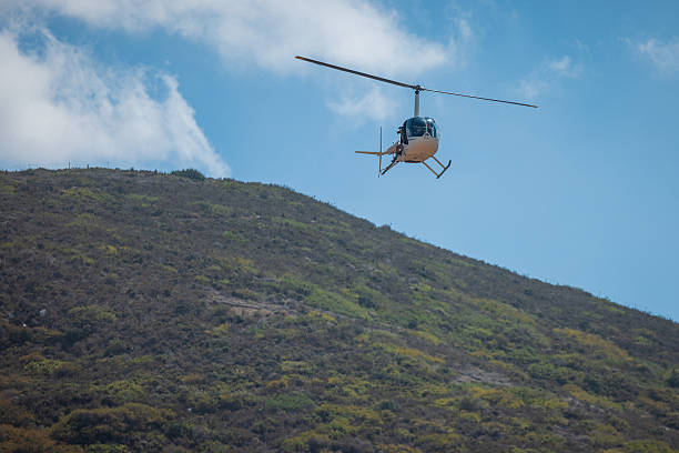A small helicopter flies above a green hillside under a clear blue sky, captured midair with wide space and natural light, conveying travel, aviation, freedom, and scenic outdoor exploration. Calm vie