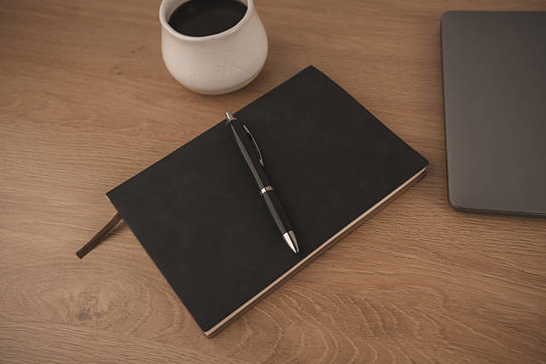 A black notebook and pen rest on a smooth marble table beside a white coffee cup filled with dark coffee. This scene captures a peaceful moment for creativity