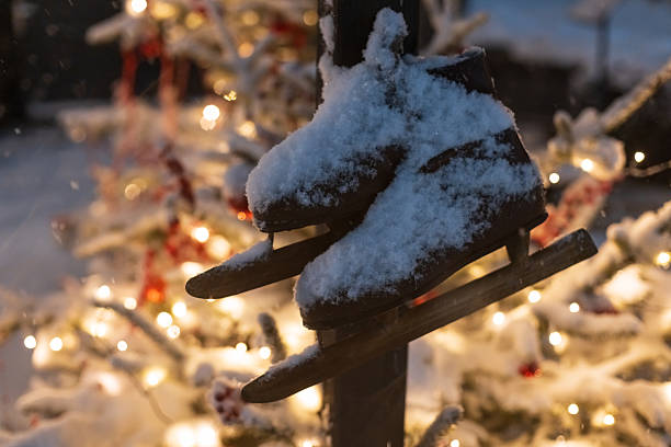Vintage ice skates covered in fresh snow hanging outdoors. A decorated Christmas tree with warm golden lights glows in the blurred background, creating a nostalgic winter holiday scene