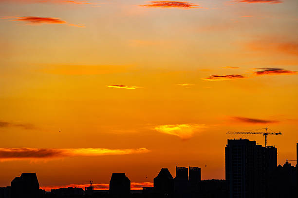 Kyiv, Ukraine. Industrial silhouettes of several heavy tower cranes on a building site in the Left Bank district against a vibrant orange sunset sky. Long lens urban landscape.