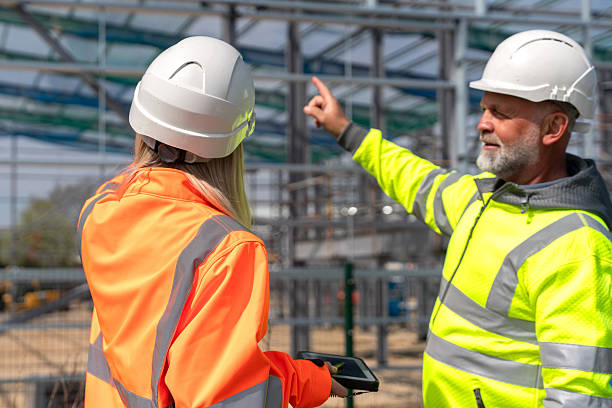 istock Two construction professionals, man and young woman, engaged in conversation at construction site, both wearing safety helmets and high-visibility jackets while reviewing plans on a tablet 2254387018