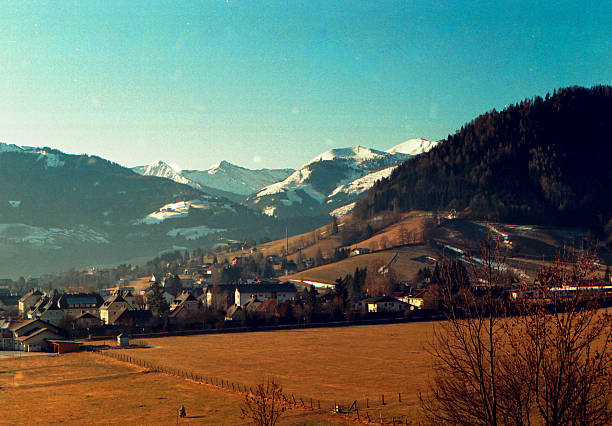 Scanned analog film photograph of traditional Alpine village nestled in valley with dramatic snow-capped peaks and warm vintage color cast