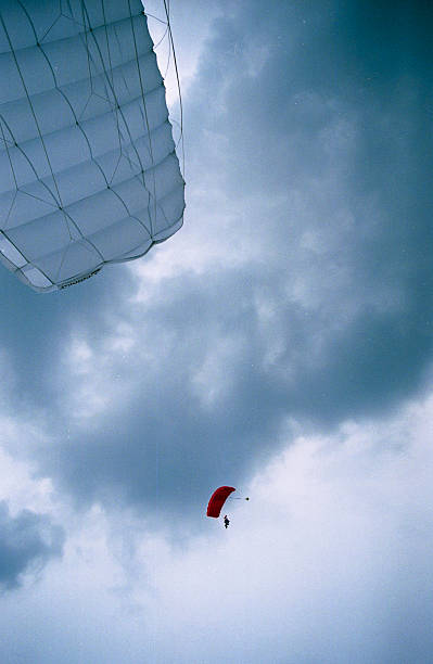 First-person perspective scanned film photograph looking up at white parachute canopy with red jumper visible against blue cloudy sky with film grain