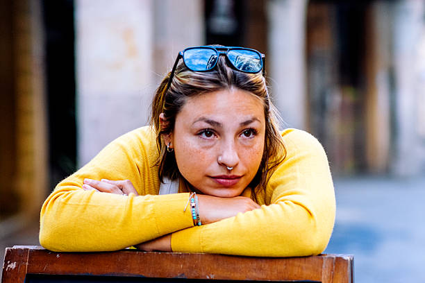 Young adult woman resting forearms on a wooden surface, wearing a yellow sweater and sunglasses on her head, looking away with a pensive expression, enjoying leisure time in an urban setting