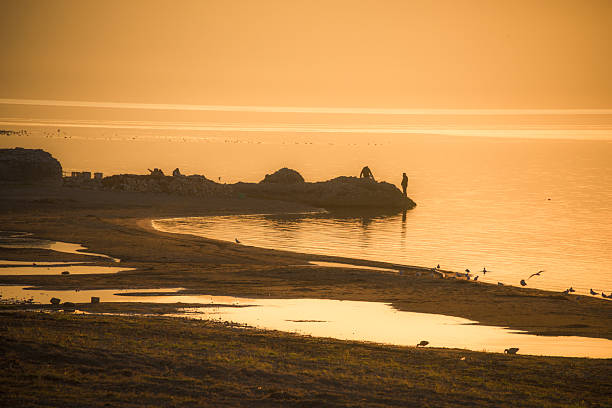 People relaxing on a sandy shoreline at golden hour, birds nearby and warm orange light reflecting on calm evening water. Iznik, bursa, turkey