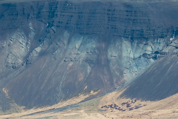 Close view of an Icelandic mountainside showing layered rock strata, steep scree slopes, and dramatic erosion patterns in muted blue-grey tones. Ideal for geology, environment, abstract nature texture, and rugged wilderness concepts.