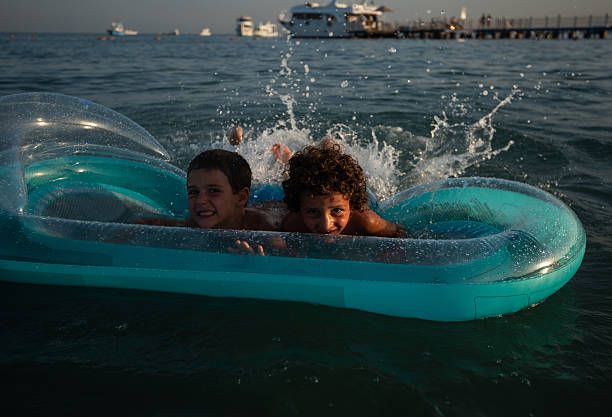 Two brothers are smiling, playing, and splashing water while floating on an inflatable raft in the mediterranean sea.