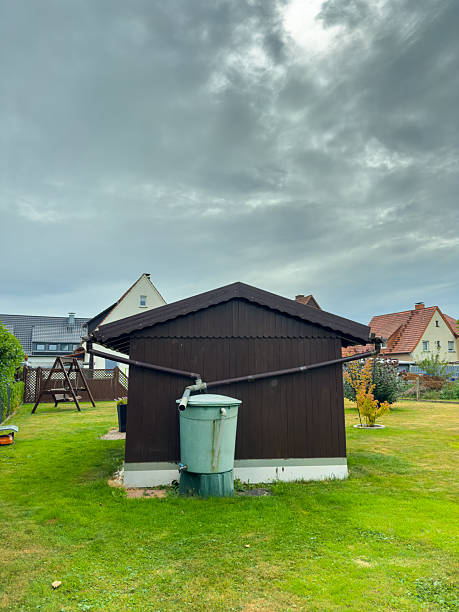 A brown garden shed stands in a lush green yard, with a water barrel nearby, under a dramatic sky. Nearby homes create a peaceful atmosphere, perfect for relaxation.