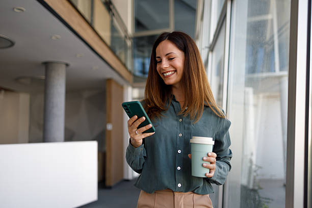 istock Professional woman smiling, using phone and holding reusable coffee cup in modern corporate office 2254404075
