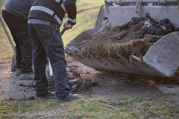 Two construction workers use shovels to load a mix of asphalt chunks and grass-covered soil into the bucket of a large excavator. The scene takes place on a roadside with green grass visible, capturing a moment of manual labor and road surface preparation. The image reflects teamwork and infrastructure maintenance.