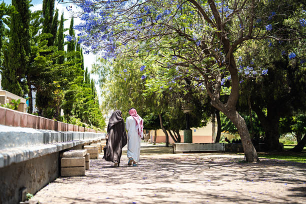 Cute Arabic couple walking through a park in Abha surrounded by blooming Jacaranda trees, enjoying a sunny day and wearing traditional attire featuring vibrant greenery and clear skies.