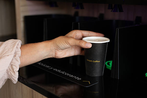 Paper cup with inscriptions of coffee and hot chocolate, placed on a black tray on a counter, with a blurred background of an illuminated coffee shop.