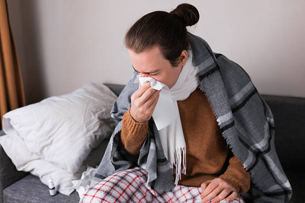 istock Sick man blowing nose into tissue while sitting on sofa wrapped in blanket. Flu, cold and allergy concept. 2254414212