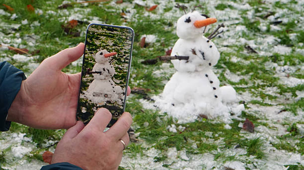 A small snowman being photographed through a smartphone on a patchy snow-covered lawn in Amsterdam. The snowman, made of two stacked snowballs, features a carrot nose, pebble eyes and buttons, and stick arms. The phone screen captures the snowman with enhanced clarity, creating a layered visual moment between reality and digital framing. The scene blends seasonal charm with modern rituals of documentation