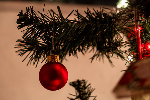 Classic red glass Christmas ornament suspended from dark green pine branch with soft background lighting. Blurred warm tones create intimate holiday atmosphere with shallow depth of field highlighting ornament detail.