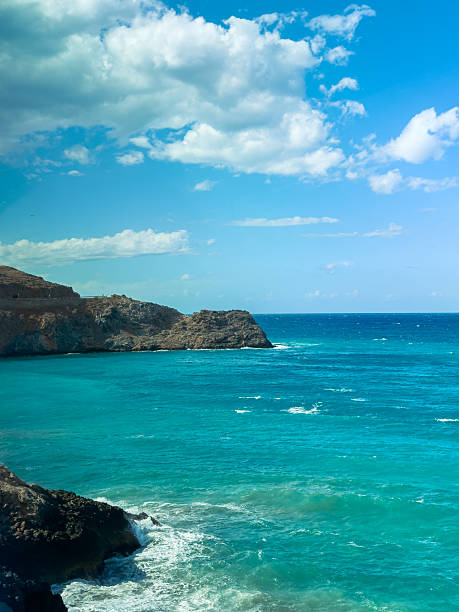 Calm turquoise waters meet the rugged coastline under a sunny sky filled with soft white clouds. The scene evokes a sense of peace and natural beauty.