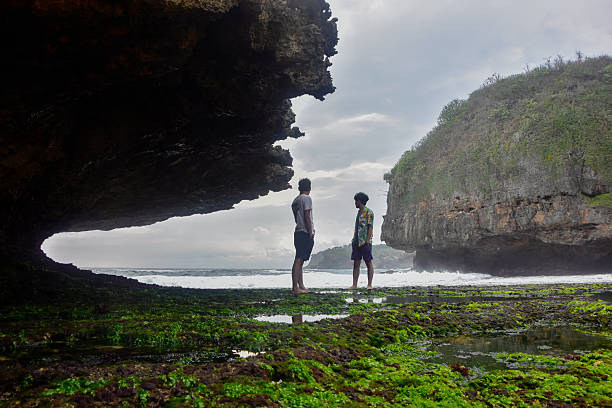 Rear view of two young Asian men standing on green moss-covered coral rocks, looking out at the moody ocean waves framed by a dramatic natural limestone cave arch in Greweng beach, Yogyakarta, Indonesia