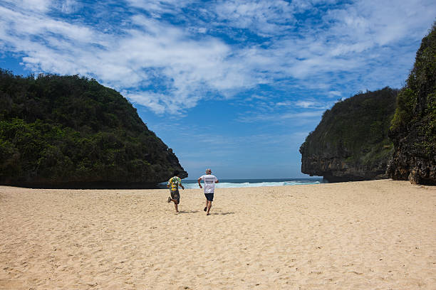 Back view of two young men running across the white sand towards the ocean waves, framed by lush green cliffs and a bright blue sky with clouds at Greweng beach, Yogyakarta, Indonesia