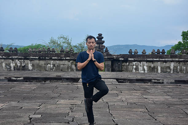 A young man in casual attire performs a yoga tree pose with hands in prayer on the stone floor of an ancient temple, symbolizing zen and mindfulness