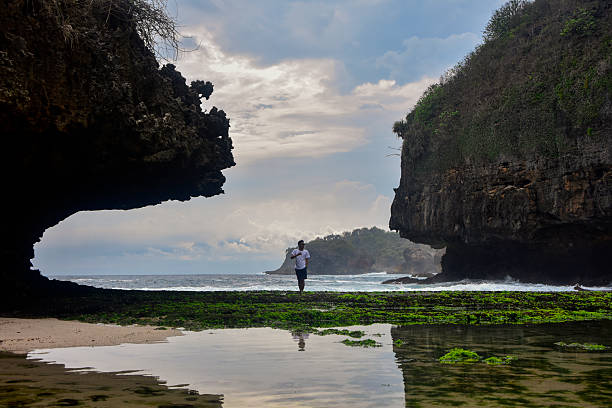 A young man stands on a vibrant green mossy shoreline framed by two massive dark rock cliffs, overlooking the blue ocean under a cloudy sky. Ideal for travel and adventure themes