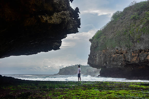 Side view of a young Asian man standing on a green algae reef during low tide, pointing his finger toward the distant ocean horizon framed by massive rock formations