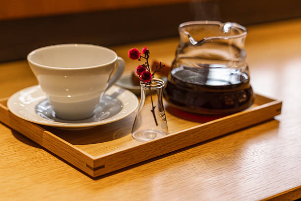 A freshly brewed coffee setup on a wooden tray, featuring a steaming glass carafe, an empty white ceramic cup, and a delicate red floral accent, creating a warm and inviting cafe ambiance for a