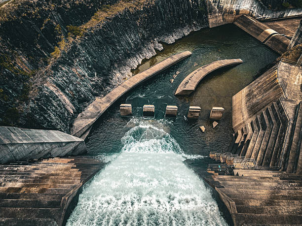 An aerial view captures the immense scale of a concrete dam, with water powerfully flowing over its tiered spillway into a churning pool below