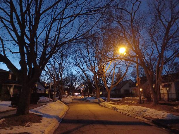 An amber streetlamp illuminates a quiet suburban street with bare trees and snow on the ground at dusk.
