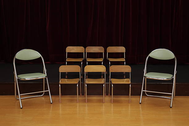 Indoor stage setup with multiple wooden chairs arranged in front of a deep red velvet curtain.