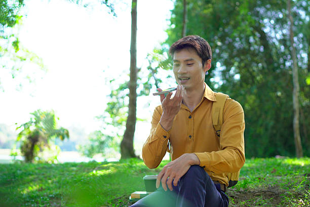 Happy asian man using a smartphone in a park, enjoying a pleasant conversation on a sunny day, surrounded by lush greenery and modern architecture..