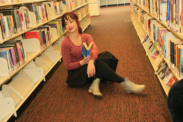 A Filipino woman sitting on the floor in a Public Library. She has long multicolored hair, makeup, a maroon top, black pants and white boots, with a book in her hand.