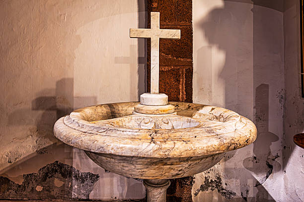 Parroquia de San Francisco de Asis, Santa Cruz, Tenerife, Spain, Europe - December 16th 2025:   A marble baptismal font with a cross, placed in a side chapel rather than in the central position commonly used in Protestant churches. The church was completed in 1640 and has served various functions over time, from monastic church to its current role as a parish church for the surrounding community. The interior decoration follows the Spanish Baroque tradition rich in gold and silver, featuring ornate sculptural details, a richly gilded altarpiece and the vertical perspective of the nave leading toward the altar.