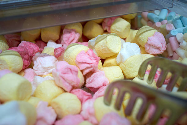 Pastel marshmallows in open bin, soft yellow pink and blue mounds with metal tongs, warm market lighting,