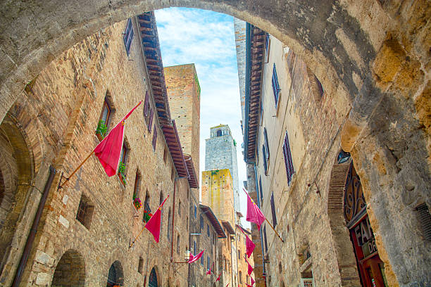 A narrow medieval street in San Gimignano, framed by a stone arch, with tall historic towers rising above warm brick buildings, Tuscany, Italy